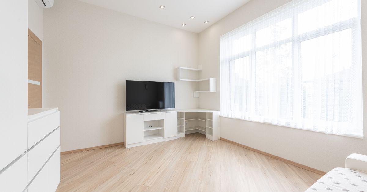 Modern TV set on cupboard placed near empty table in spacious room with big window and white cabinets in apartment